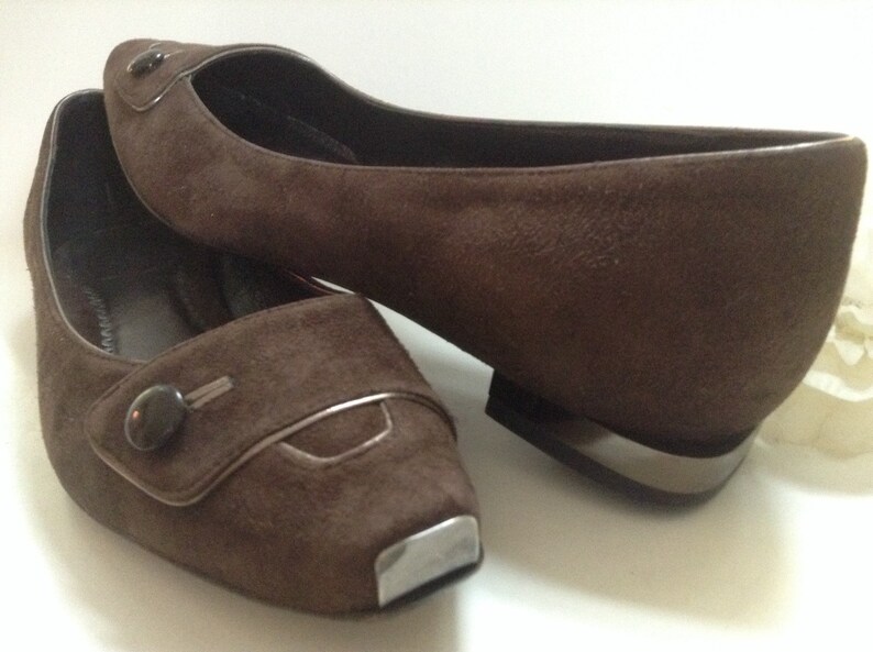 May include: A pair of brown suede flats with a decorative button on the toe. The shoes have a silver trim around the toe and heel.