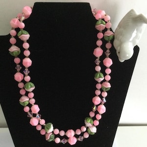 May include: A double strand pink and green glass bead necklace with a floral design. The necklace is displayed on a black stand.