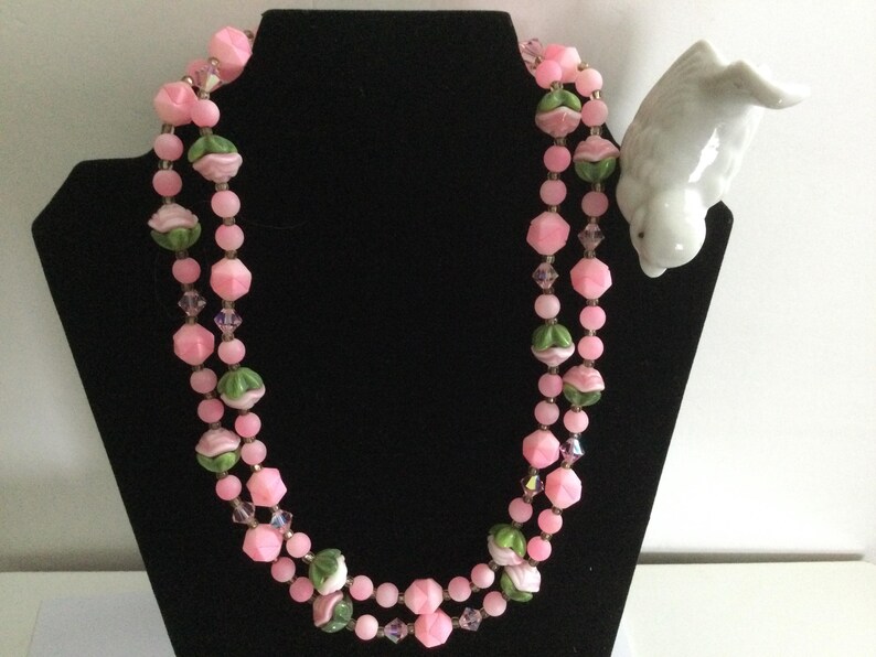 May include: A double strand pink and green glass bead necklace with a floral design. The necklace is displayed on a black stand.