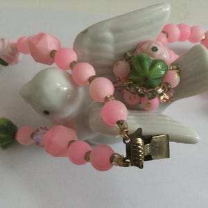 May include: A pink and green beaded necklace with a floral design and a metal clasp. The necklace is displayed on a white ceramic bird figurine.