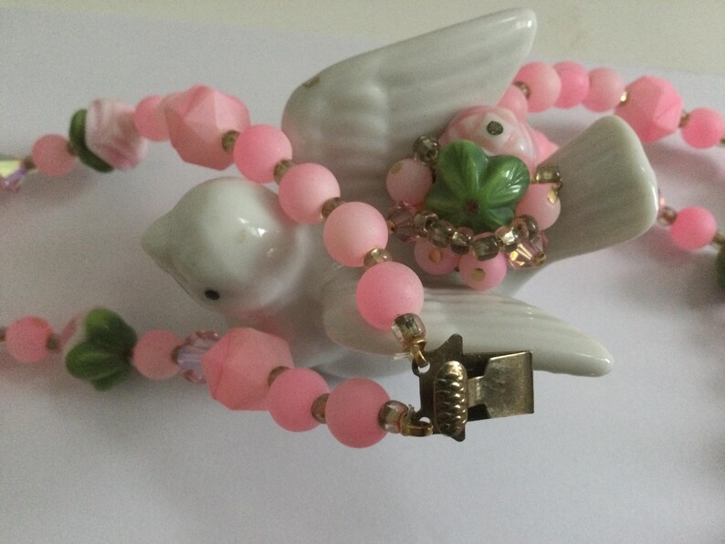 May include: A pink and green beaded necklace with a floral design and a metal clasp. The necklace is displayed on a white ceramic bird figurine.