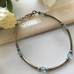 May include: A silver bracelet with a clasp and blue glass beads. The bracelet is on a white surface with the text "PLATA DENLEY" visible.