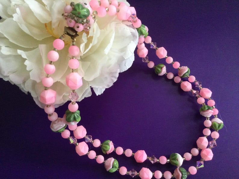 May include: A double strand pink and green glass bead necklace with flower accents. The necklace is on a purple background.