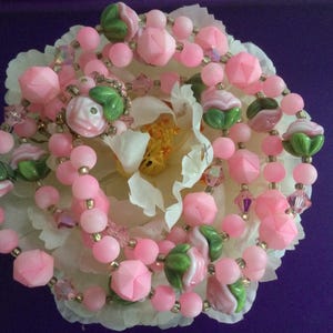 May include: A pink and green beaded necklace with flower-shaped beads. The necklace is on a white flower.