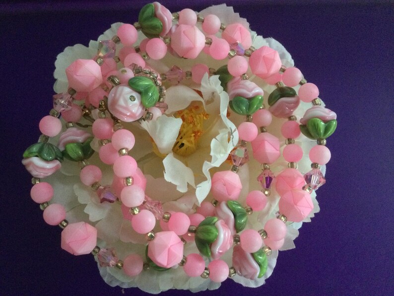 May include: A pink and green beaded necklace with flower-shaped beads. The necklace is on a white flower.