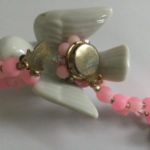 May include: A pink and white beaded necklace with a porcelain dove charm. The dove has a gold-tone button on its chest and a small gold-tone tag on its wing.