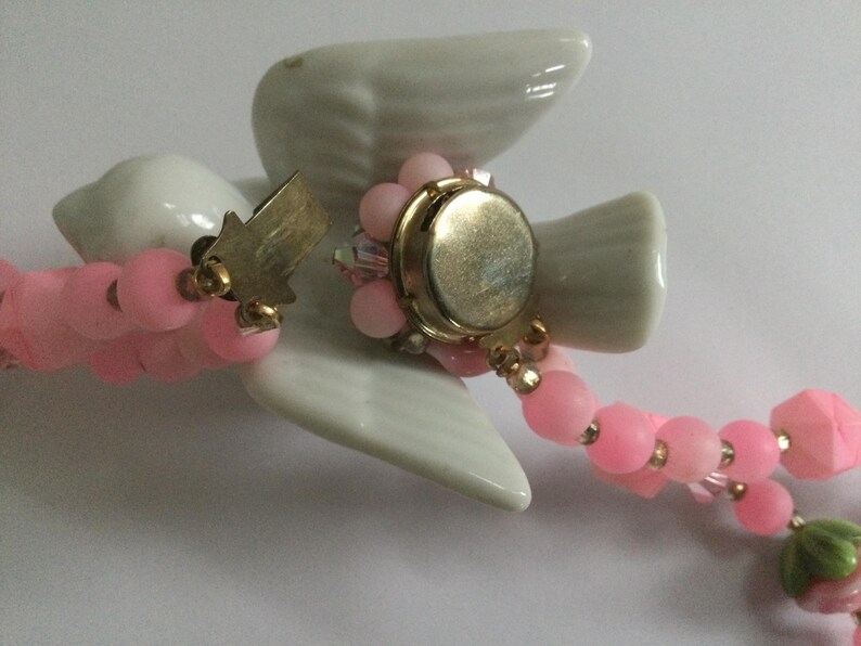 May include: A pink and white beaded necklace with a porcelain dove charm. The dove has a gold-tone button on its chest and a small gold-tone tag on its wing.