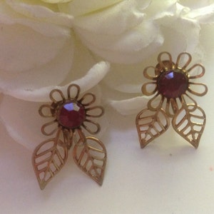 Op de afbeelding: Een paar gouden oorbellen met een rood bloemenontwerp. Elke oorbel heeft een rode edelsteen in het midden van de bloem en twee bladeren.