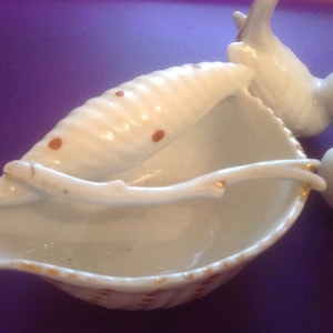 Puede incluir: Plato de porcelana blanca en forma de concha de mar con un peque&ntilde;o p&aacute;jaro blanco posado en el borde. El plato est&aacute; decorado con lunares marrones y tiene un borde dorado.