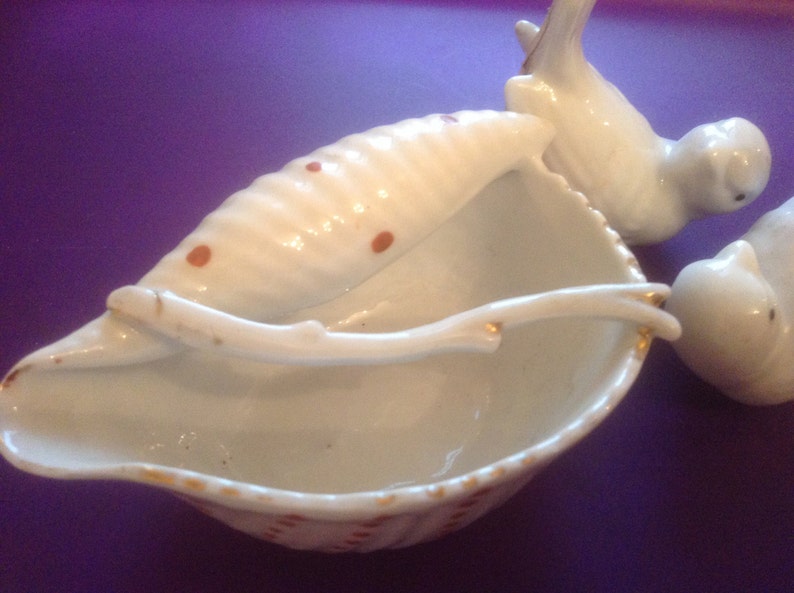 Puede incluir: Plato de porcelana blanca en forma de concha de mar con un peque&ntilde;o p&aacute;jaro blanco posado en el borde. El plato est&aacute; decorado con lunares marrones y tiene un borde dorado.