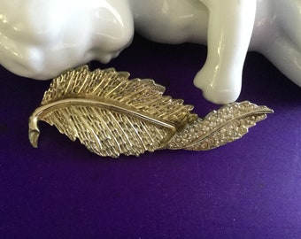 Op de afbeelding: Een goudkleurige blad-vormige broche met ingewikkelde details. Eén kant van het blad is getextureerd, terwijl de andere kant is versierd met kleine, sprankelende strass-steentjes. De broche ligt op een paarse achtergrond.