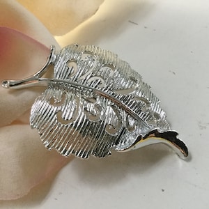 Op de afbeelding: Een zilveren broche in de vorm van een blad met een delicaat, ingewikkeld ontwerp. De broche heeft een getextureerd oppervlak en een uitgesneden patroon.