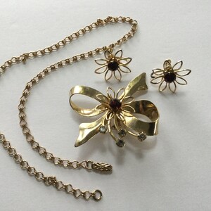 May include: A gold tone necklace with a bow brooch and matching earrings. The brooch features a large red rhinestone in the center of a flower design. The earrings are also flower shaped with red rhinestones in the center.