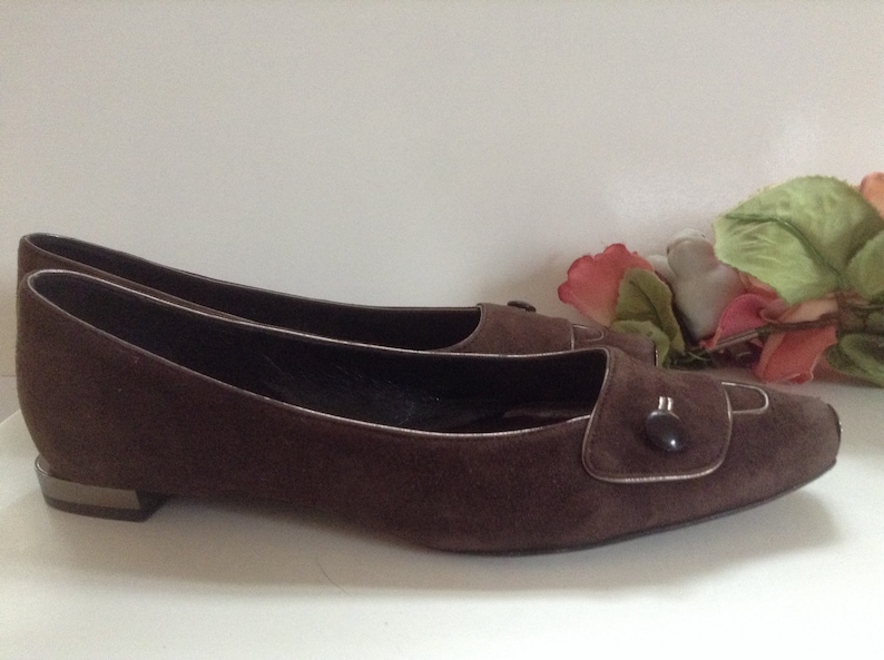 May include: Brown suede ballet flats with a button detail on the toe. The flats have a pointed toe and a low heel.