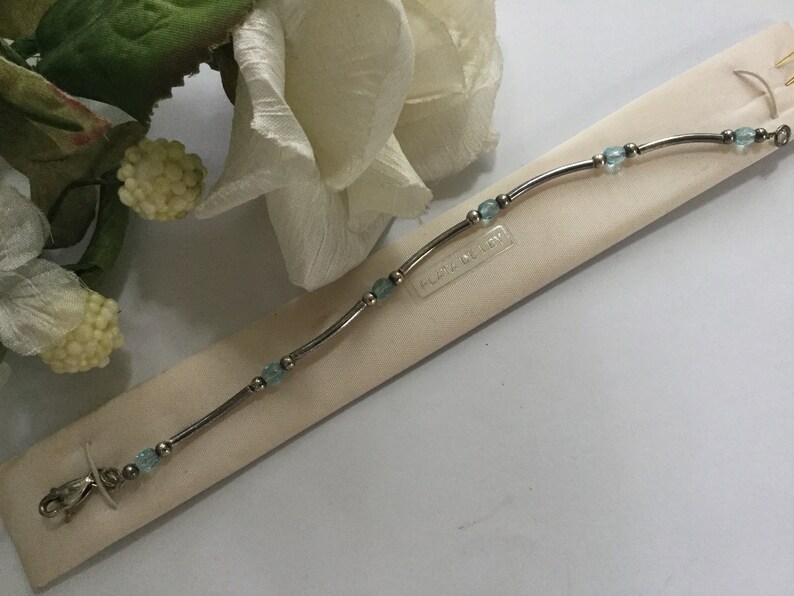 May include: A silver bracelet with light blue beads and a clasp. The bracelet is on a white background with the text "Flora & Joy" printed on it.