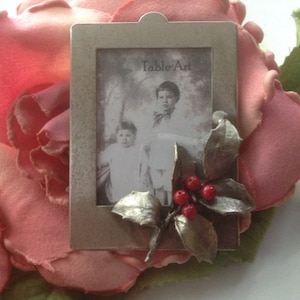 May include: A silver-toned picture frame with a black and white photo of two children. The frame is decorated with a holly sprig and red berries. The frame has the text "Table Art" printed on it.