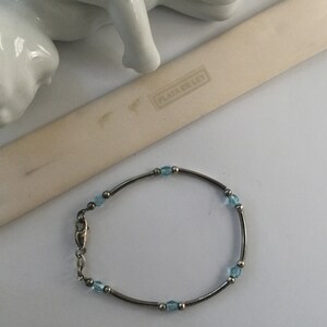 May include: A silver bracelet with light blue beads and a clasp. The bracelet is on a white surface with a small white figurine and a white card with the text "PLATA DE LEY" in black.