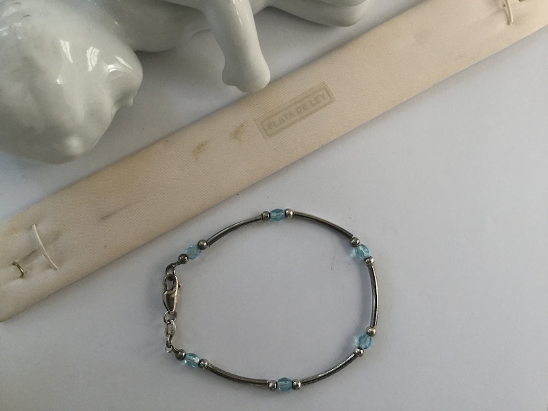 May include: A silver bracelet with light blue beads and a clasp. The bracelet is on a white surface with a small white figurine and a white card with the text "PLATA DE LEY" in black.