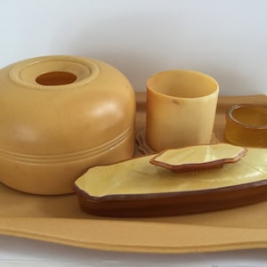 May include: A vintage vanity set with a round, cream-colored lidded jar, a small cream-colored cup, a small amber-colored jar, and a rectangular cream-colored tray.