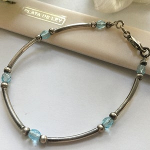 May include: A silver bracelet with a clasp closure and blue gemstone beads. The bracelet is on a white surface with the text "PLATA DE LEY" visible.
