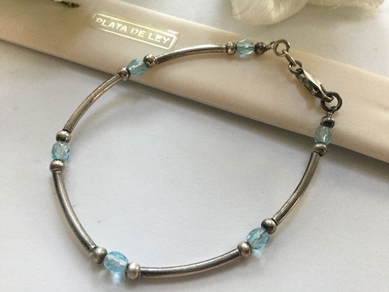 May include: A silver bracelet with a clasp closure and blue gemstone beads. The bracelet is on a white surface with the text "PLATA DE LEY" visible.