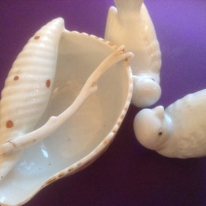 Puede incluir: Un conjunto de tres figuritas de p&aacute;jaros de porcelana blanca. Un p&aacute;jaro est&aacute; posado en un plato con forma de concha con un borde dorado. La concha tiene manchas marrones y un mango de rama dorada.