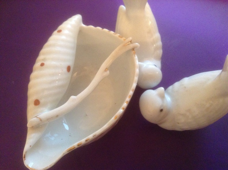 Puede incluir: Un conjunto de tres figuritas de p&aacute;jaros de porcelana blanca. Un p&aacute;jaro est&aacute; posado en un plato con forma de concha con un borde dorado. La concha tiene manchas marrones y un mango de rama dorada.