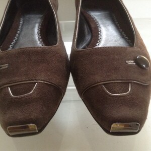 May include: A pair of brown suede flats with a silver trim and a button detail. The shoes have a pointed toe and a low heel.