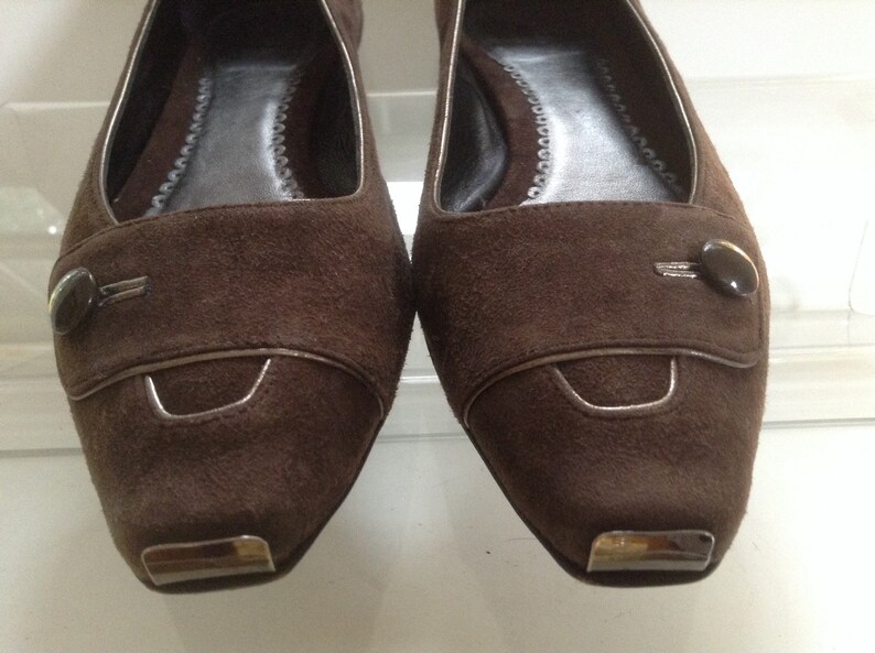 May include: A pair of brown suede flats with a silver trim and a button detail. The shoes have a pointed toe and a low heel.