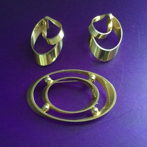 May include: A set of three gold-toned jewelry pieces. Two pieces are earrings with a simple oval shape and a smaller oval shape inside. The third piece is a brooch with a larger oval shape and a smaller oval shape inside. The brooch has four small gold balls on the outer edge.