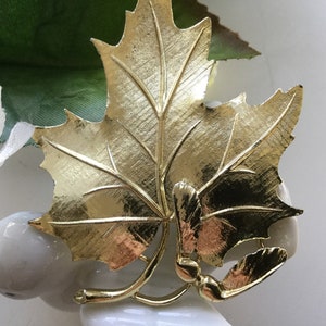 May include: A gold-toned maple leaf brooch with a detailed design. The leaf is textured and has a stem with a small seed pod.