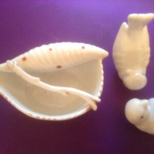 Puede incluir: Tres figuritas de cer&aacute;mica blanca, dos p&aacute;jaros y un plato con forma de concha de mar con manchas marrones. Los p&aacute;jaros est&aacute;n de pie y el plato con forma de concha de mar est&aacute; abierto.