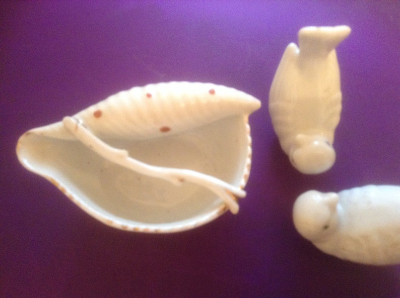 Puede incluir: Tres figuritas de cer&aacute;mica blanca, dos p&aacute;jaros y un plato con forma de concha de mar con manchas marrones. Los p&aacute;jaros est&aacute;n de pie y el plato con forma de concha de mar est&aacute; abierto.