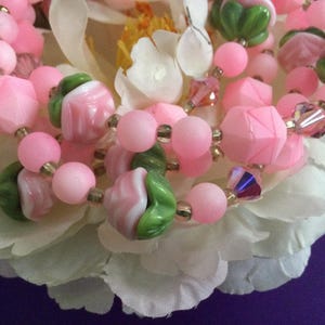 May include: A close-up of a pink and green beaded necklace. The beads are made of glass and have a variety of shapes and sizes. The necklace is lying on a white flower.