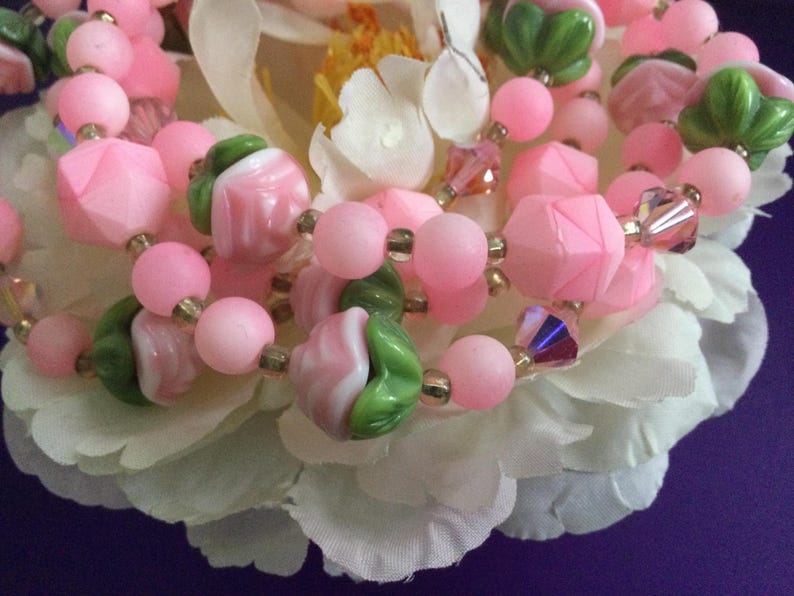 May include: A close-up of a pink and green beaded necklace. The beads are made of glass and have a variety of shapes and sizes. The necklace is lying on a white flower.
