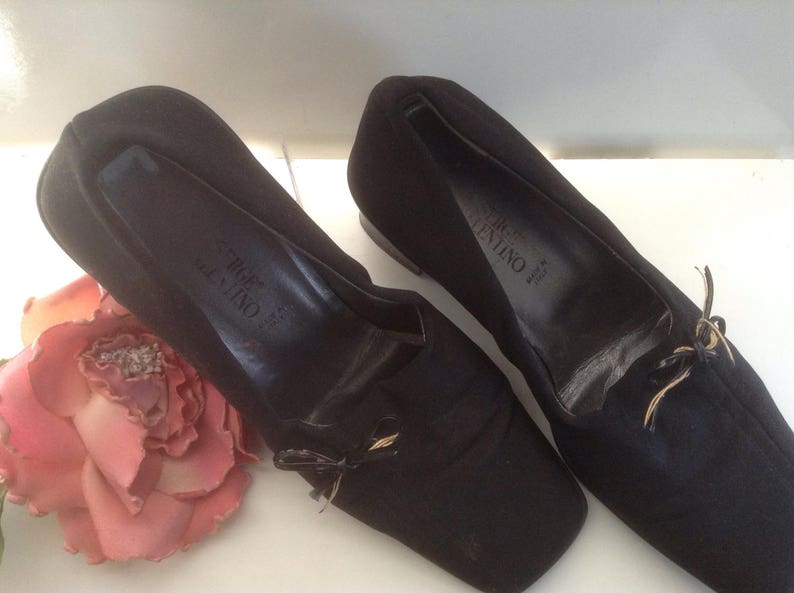 May include: A pair of black satin Valentino flats with a gold bow detail. The shoes have a pointed toe and a low heel.