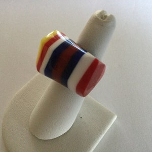 May include: A colorful, multi-striped ring made of glass. The ring is red, white, blue, and yellow. It is displayed on a white ring holder.
