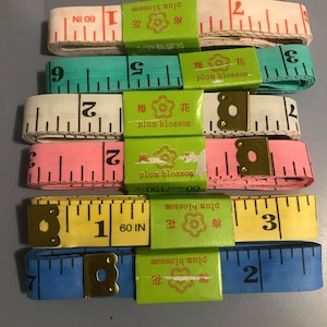 May include: A set of six colourful measuring tapes with a floral design and the text "plum blossom" in English and Chinese. The tapes are in various colours, including blue, yellow, pink, and white. Each tape has a brass end cap and measures 60 inches (152.4 cm).