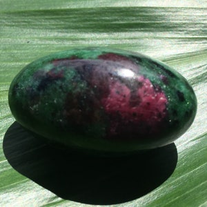以下が含まれることがあります： 緑と深紅の色合いが混ざった、楕円形の研磨された石。石は滑らかで反射性があり、緑の葉の上に置かれています。色は混ざり合い、ユニークなパターンを作り出しています。石の形は細長く丸みを帯びています。