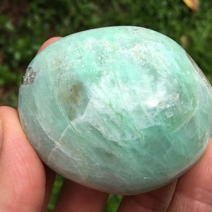 Può includere: Una pietra naturale liscia, verde e lucidata con un motivo marmoreo verde chiaro e bianco. La pietra ha una forma ovale e una finitura lucida.