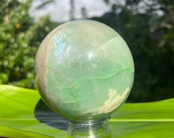 May include: A green and white crystal sphere on a clear stand. The sphere has a smooth, polished surface and is made of natural stone.