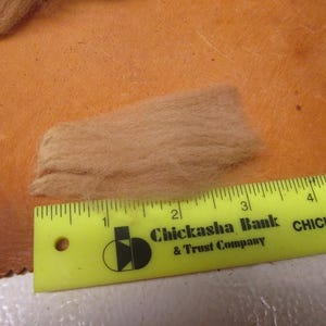 May include: A close-up of a small pile of light brown wool fibers next to a yellow ruler with the text "Chickasha Bank & Trust Company" and "CHICKASHA, OK".
