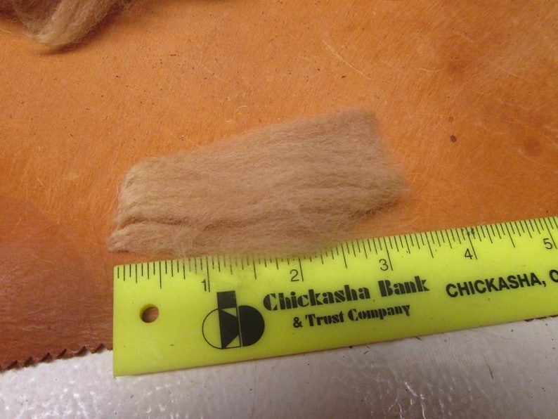 May include: A close-up of a small pile of light brown wool fibers next to a yellow ruler with the text "Chickasha Bank & Trust Company" and "CHICKASHA, OK".