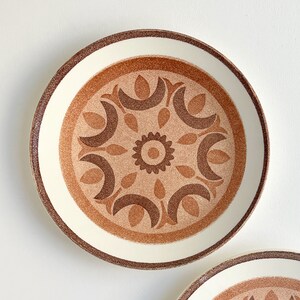 May include: A white ceramic plate with a brown and tan geometric pattern. The pattern features a central flower with petals and leaves.