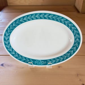 May include: White oval serving platter with a teal border featuring a leaf and floral design. The platter is supported by a clear plastic stand and is set against a wooden background. A decorative piece for serving food.