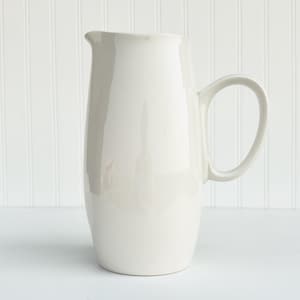 May include: A white ceramic pitcher with a wide base and a rounded body. The pitcher has a handle and a smooth, glossy finish.