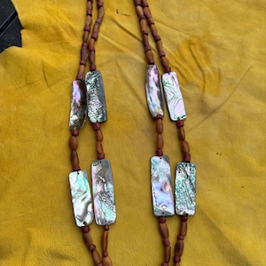 May include: A long necklace featuring two strands of brown wooden beads interspersed with small red beads and rectangular, iridescent abalone shell segments. The necklace is constructed with a leather cord at the top, showcasing natural textures and varied shell colors.