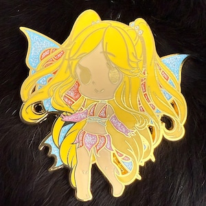 May include: A gold-coloured enamel pin featuring a cartoon fairy with long blonde hair, wearing a pink and purple outfit and blue wings with glitter accents.