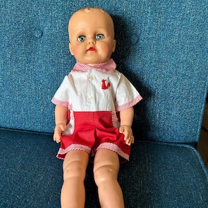 May include: A vintage baby doll with blue eyes, red lips, and a bald head. The doll wears a white shirt with red trim and a red anchor emblem, paired with red shorts. The doll is seated on a blue upholstered surface.