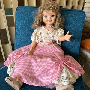 May include: A vintage doll with blonde curly hair, wearing a white lace top and a pink gingham dress. The doll is seated on a blue chair, with a pearl necklace and a pink bow in her hair. The doll's shoes are beige.
